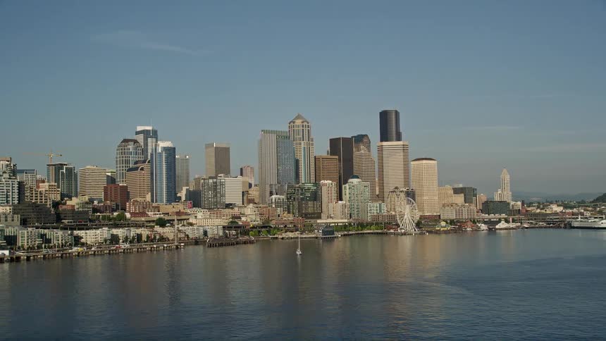  aerial footage of downtown Seattle's Central Waterfront showcasing the city's skyline busy metropolis and iconic Ferris wheel with the shoreline of Elliott Bay in the backdrop on a sunny day