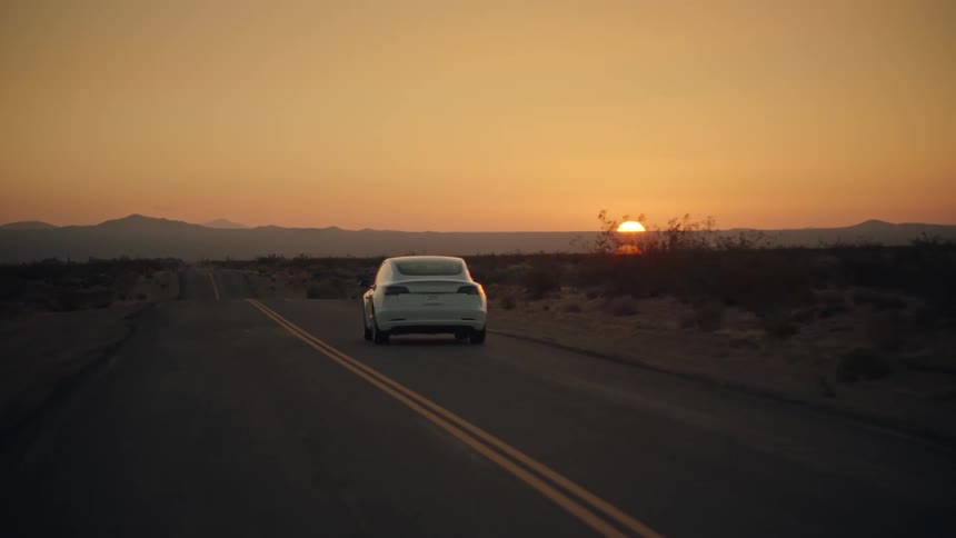 A white automobile travels on a desert road as the sun sets over a mountain in the distance under a hazy orange sky