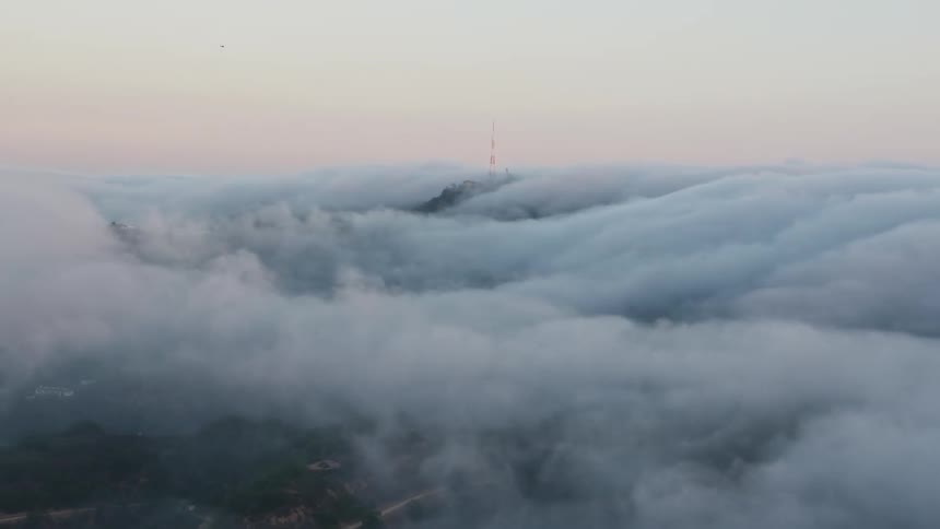Aerial 4K footage of a radio tower standing tall amidst a thick marine layer with the sunset and Hollywood Hills in the background in Los Angeles