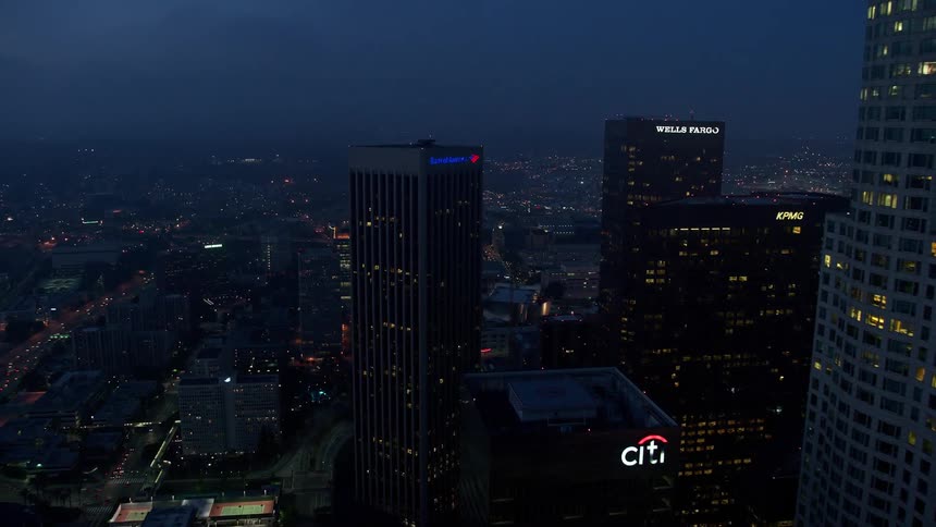 An aerial view of multiple skyscrapers with illuminated bank signs in downtown Los Angeles during twilight