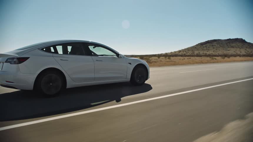 The sun shines brightly upon a white automobile as it travels on a desert road and there is a mountain in the distance