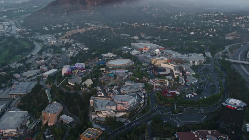 Approaching aerial view of Universal Studios Hollywood at dusk with partly cloudy skies and misty hills in the background