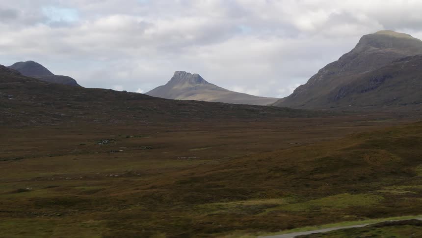 A dark sports car travels on a rural road near mountains and there are mountains in the distance under a cloudy sky
