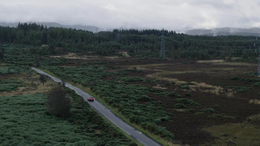 Utility towers stretch cables across a rural area as an automobile travels on the road toward forests and there are mountains in the distance