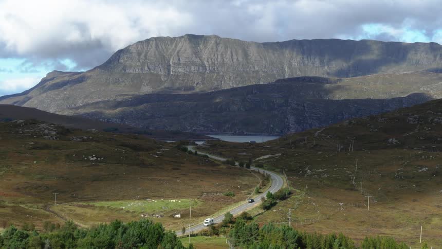 An aerial view of a road through grassy fields leading toward water at the foot of a mountain