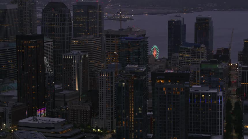 A skyscraper in the center of a tower block of a coastal city at dusk with many office lights turned on