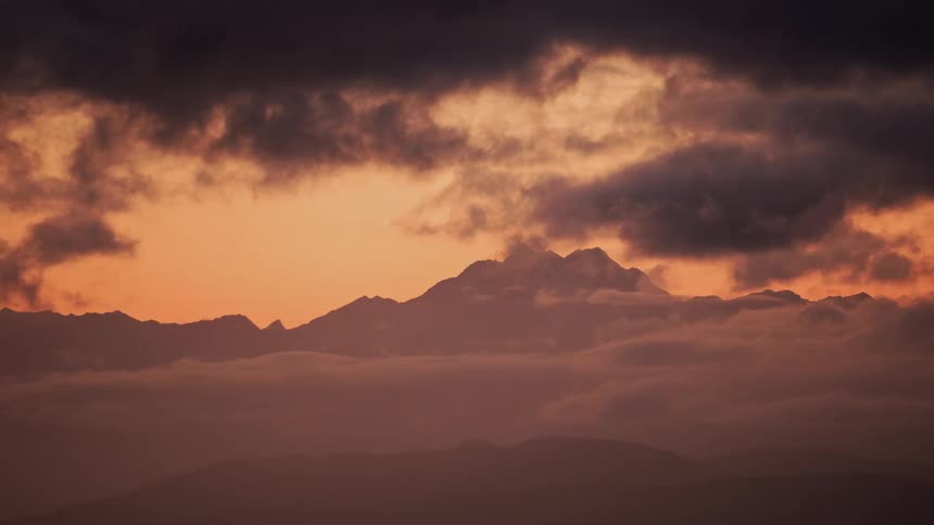 Clouds float across the sky under orange light from the setting sun with mountains in the distance