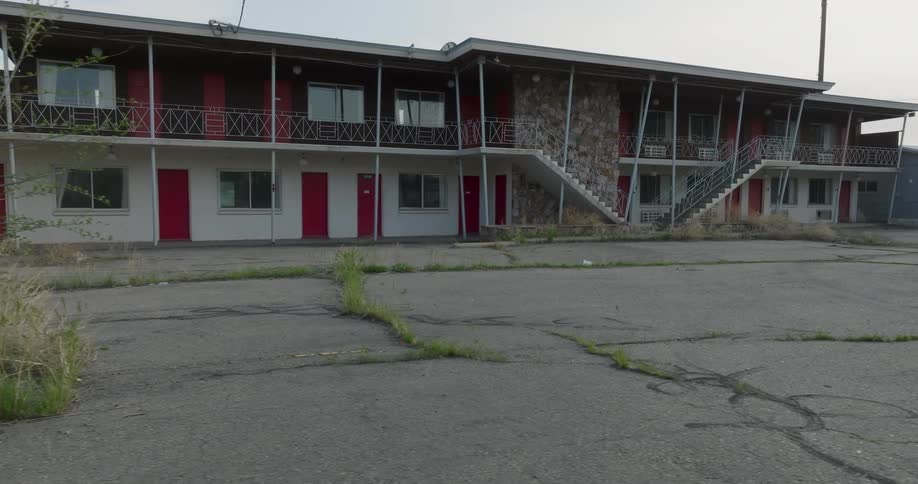 Revealing the front of an old motel from behind a bush growing in cracks of the parking lot