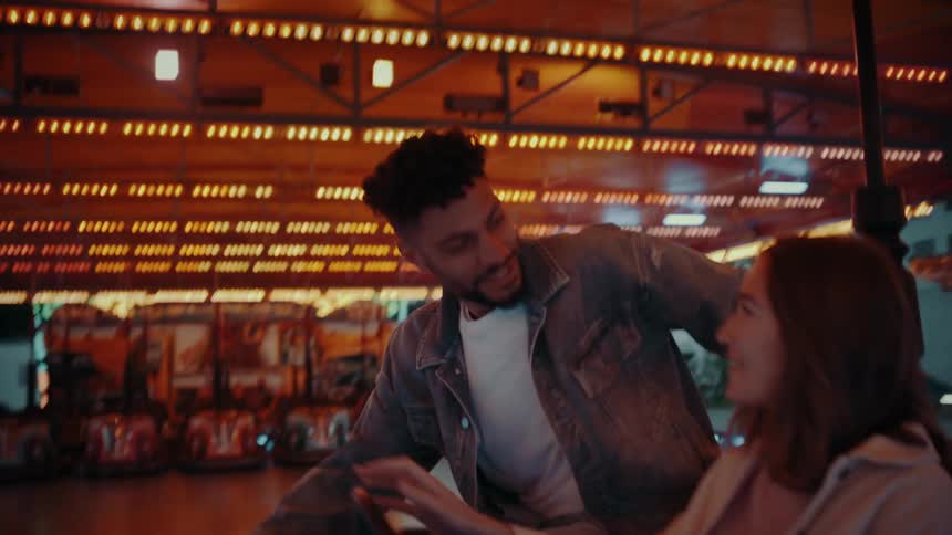 Young man steps into a bumper car to join a young woman on a bumper car ride they are both smiling and he puts his arm around her