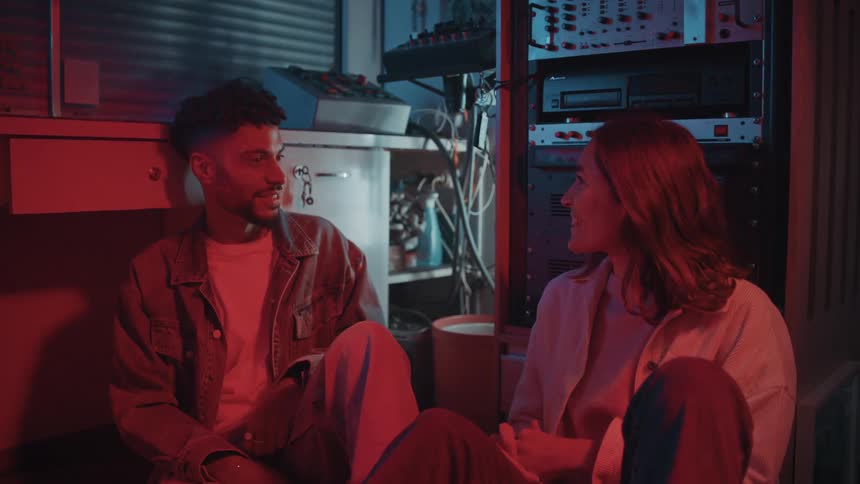 A man and woman sit on a floor and talk in a room full of audio equipment and mixers