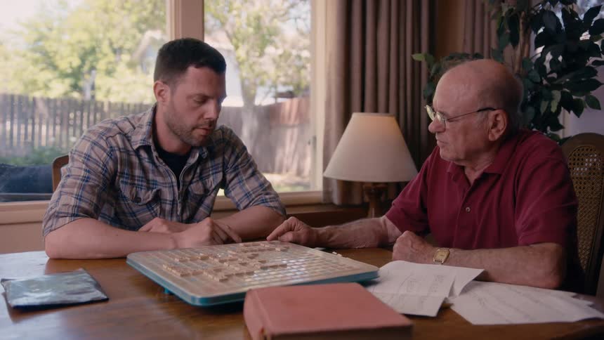 Two men one of them elderly play scrabble together in a living room