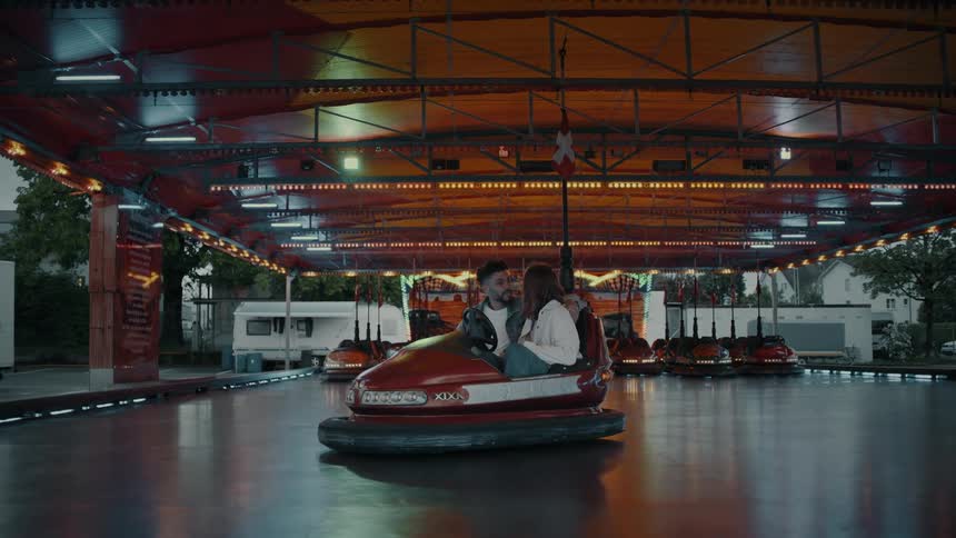 Brown curly haired man wearing denim and redhead woman wearing a white jacket chat on a red bumper car