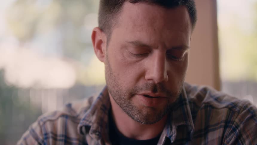 Man with stubble beard and plaid shirt talking to person sitting at table