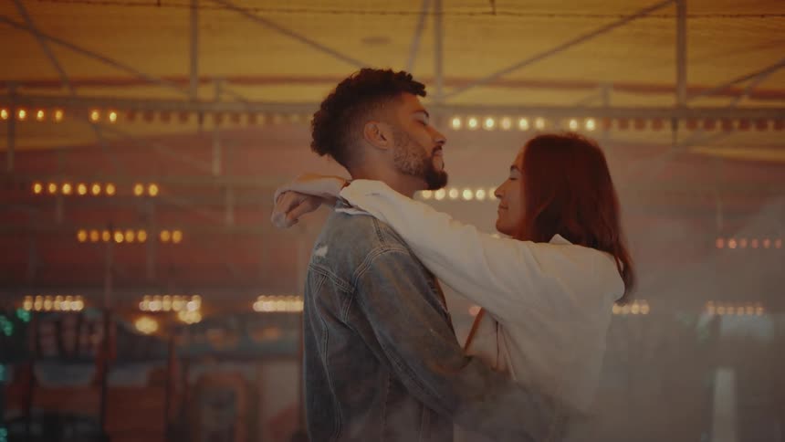 A man and a woman slow dance in front of a carousel as smoke covers them