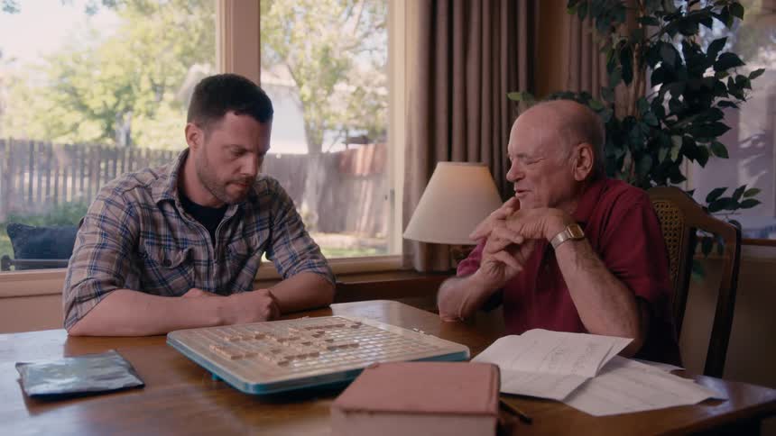 Two men are playing scrabble and talking to each other with the oldest of them smiling and rubbing his hands while the other remains focused on the game board