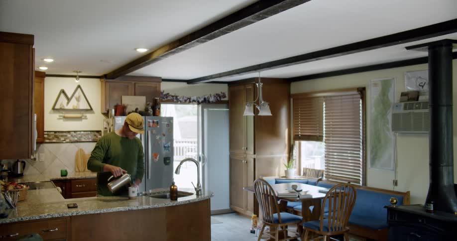 A man standing at his kitchen counter pours hot water into a mug and opens a cabinet