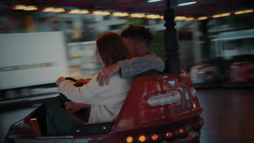 A couple driving in bumper car his arm over her shoulder and both laughing