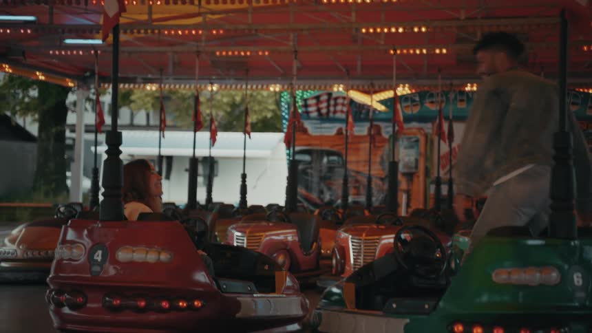 Joyous brown man wearing denim and white redhead woman in a white coat play around with bumper cars