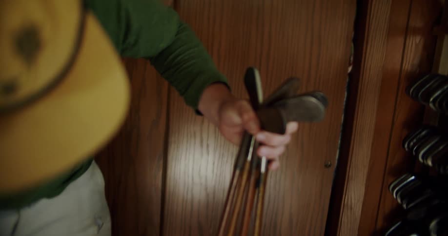 Interior shot of a man wearing a green sweater and a cap lifting up a golf bag and handling the clubs