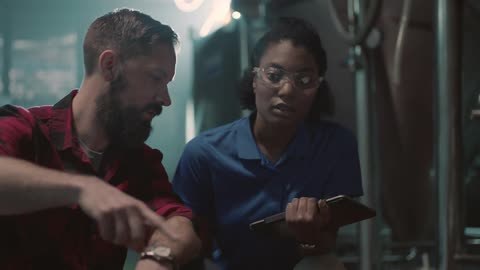 A man and woman are sitting near machinery and discussing the machinery