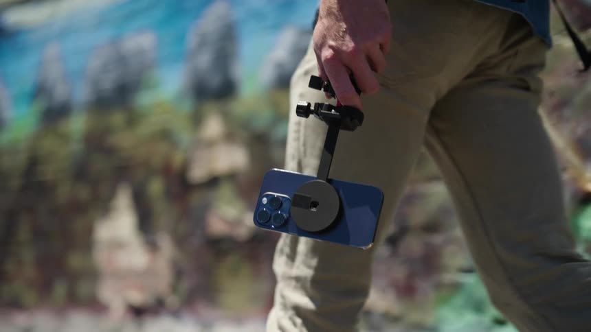 A man is holding his cell phone on a selfie stick as he walks outside near a mural painted on a wall