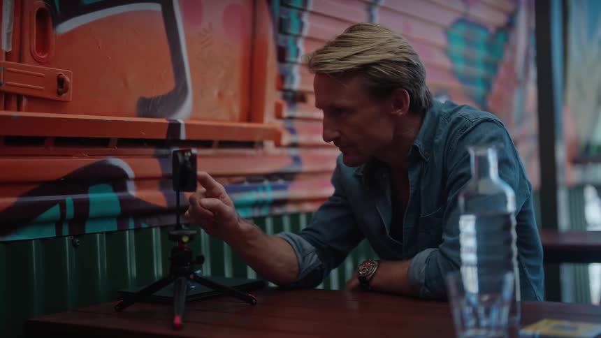 Blonde man with blue shirt sits at table using smartphone next to wall with grafitti