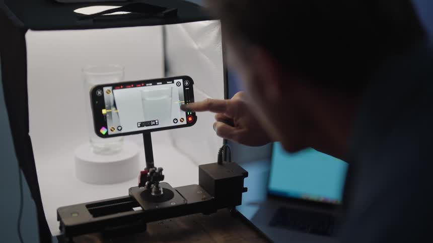 A man is taking a picture of a glass inside of a machine with his cell phone on a moving stand as he presses buttons on his phone