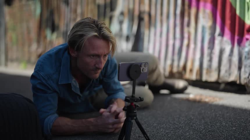 Man with blue shirt and brown pants lies on the ground using smartphone on tripod