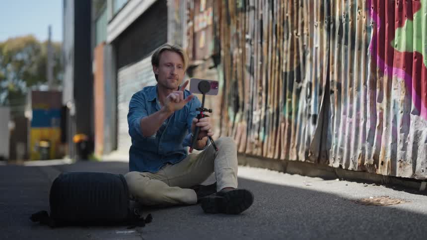 A man is sitting in an alley holding his cell phone on a small tripod and he grabs his backpack as he stands up and walks away