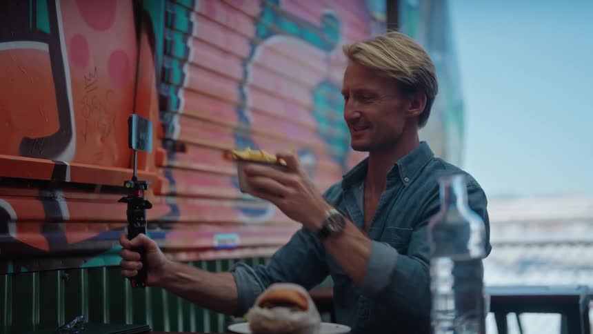 Man with blue shirt sitting at table films himself and showing a bowl of french fries at restaurant