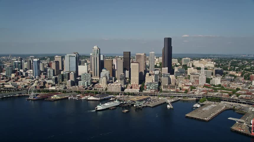 Aerial footage approaching Downtown Seattle skyline and flying over the Seattle Ferry Terminal on the Waterfront towards Central Seattle Washington showcasing the high-rise buildings wharf and bustling city life with boats in the water and mountains in th