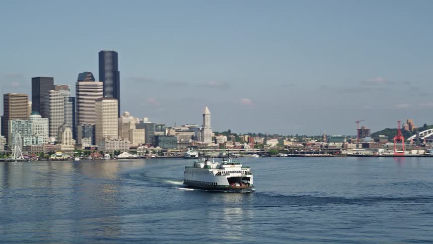 A low-flying aerial shot tracks a ferry crossing Elliott Bay revealing the downtown Seattle skyline with towering buildings and boats passing by
