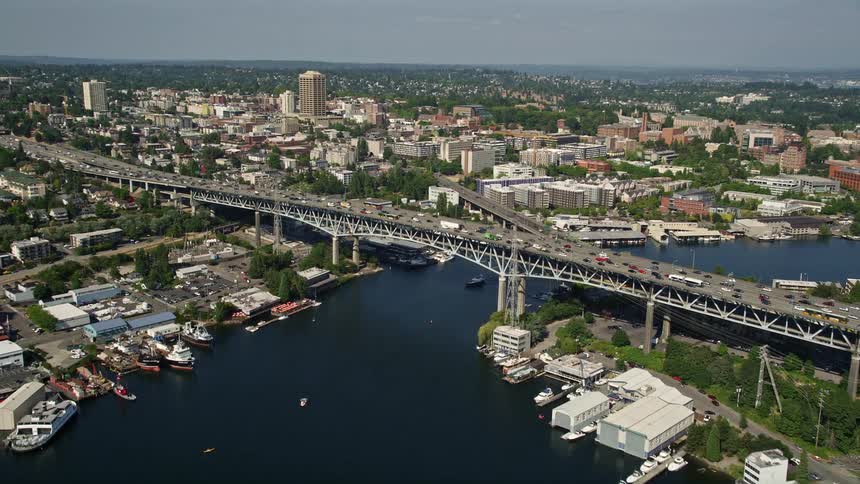 Aerial footage of the Ship Canal Bridge in Seattle Washington as traffic crosses the bridge near boat docks and continues towards downtown with a sunny day and beautiful shoreline view
