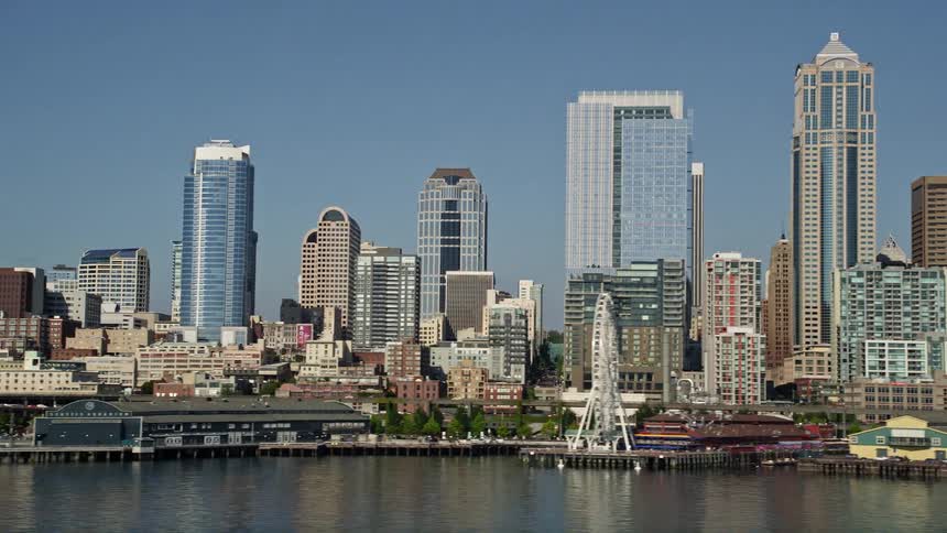 Aerial footage of the Downtown Seattle skyline and Central Waterfront viewed from Elliott Bay during a sunny day in Washington showcasing the high-rise buildings wharfs and cityscape of this bustling metropolis