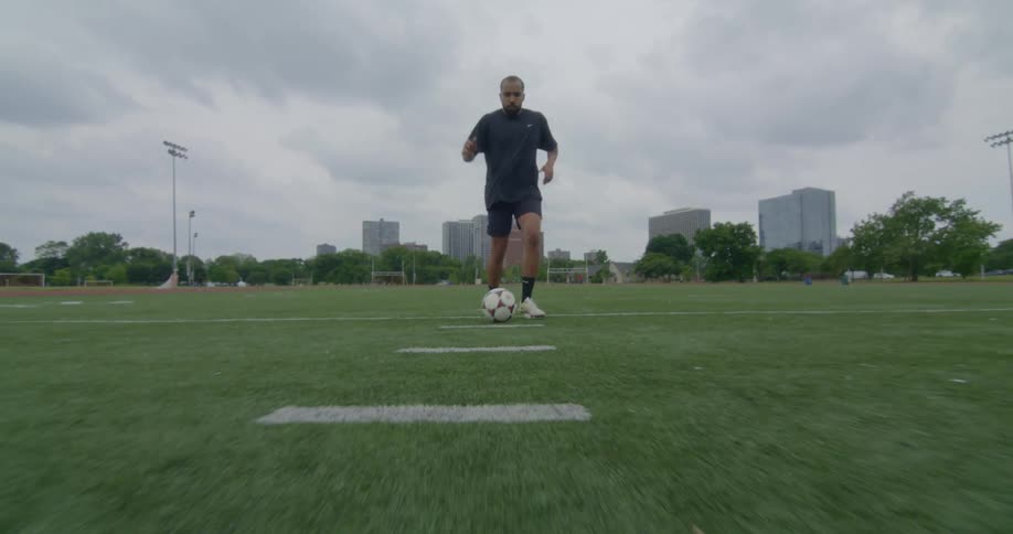 Man runs and dribbles a soccer ball in field with traffic passing on nearby street in the background