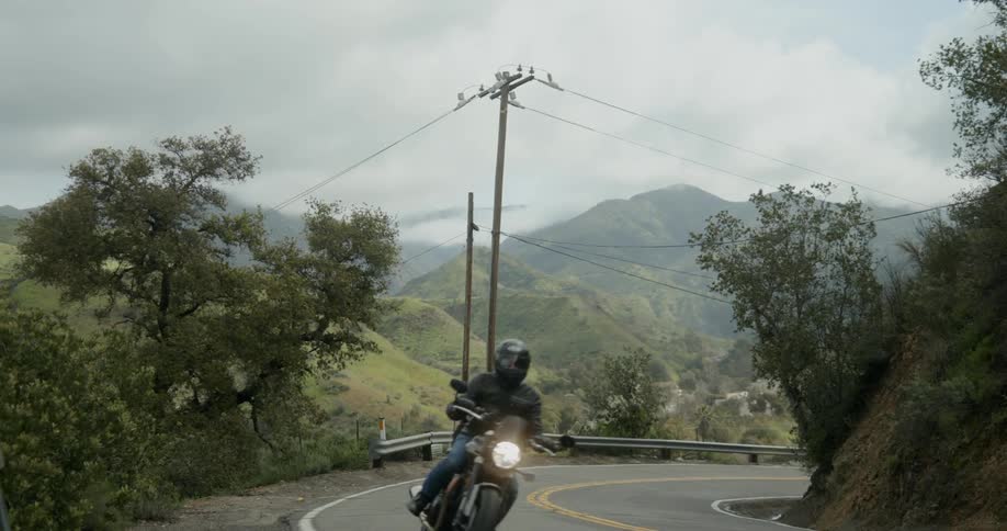 Two motorcyclists drive around highway curve with foggy mountains in the background