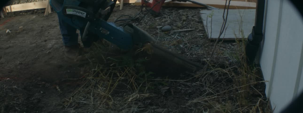 Worker uses chainsaw to cut into ground near building a short distance from wooden frame for walkway
