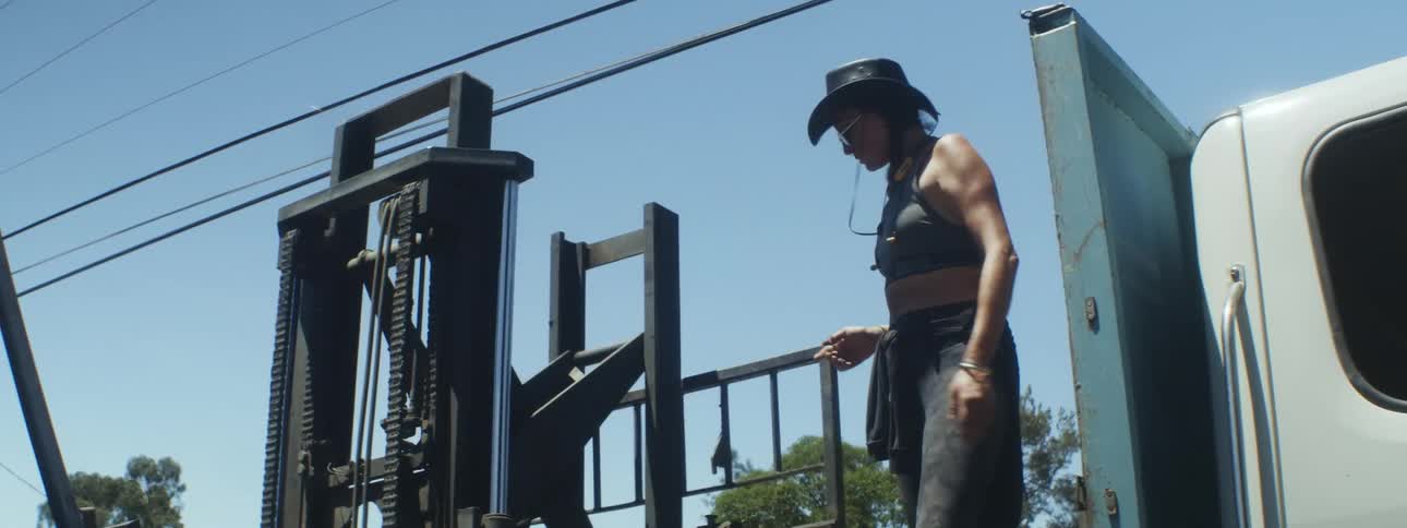 Woman stands on bed of truck and gives instructions as robotic arm picks up stones then she gets down