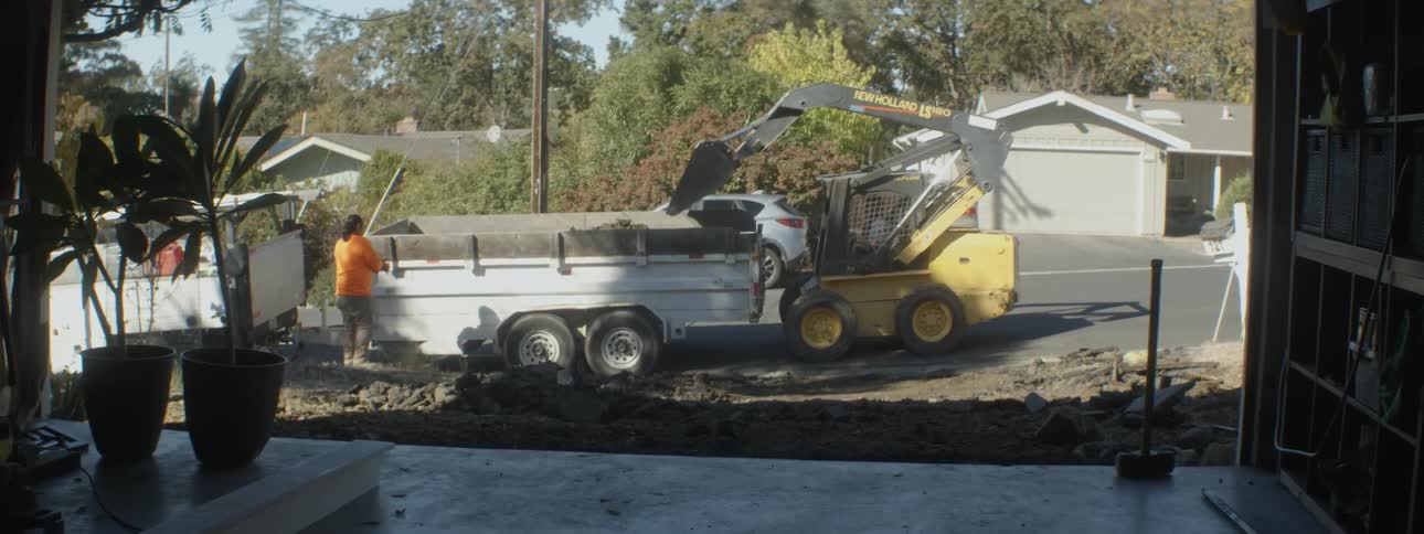 A digger machine collects dirt from a suburban street and places it in the back of a truck