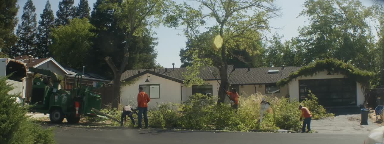Several men in orange shirts doing yard work in front of a house including tree trimming and leaf blowing midday