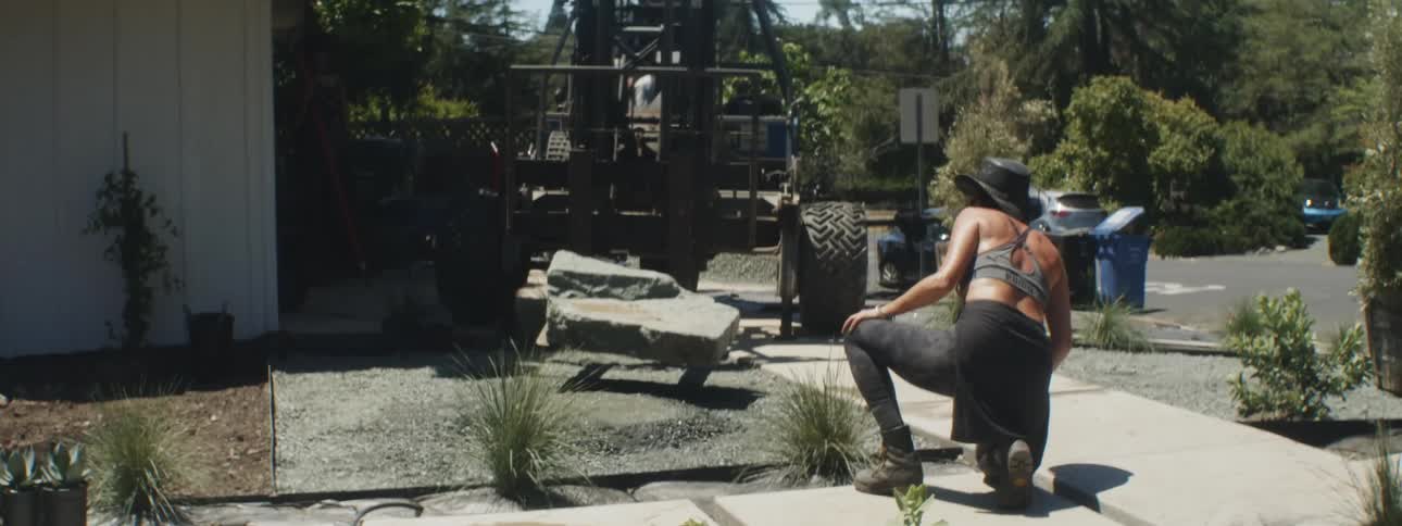 Woman kneels while watching forklift place large stone onto gravel in her front yard