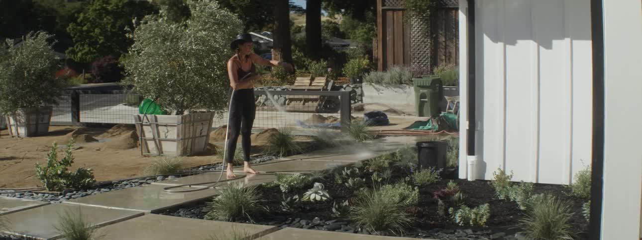 Woman stands barefoot on sidewalk and waters garden as she points out different plants