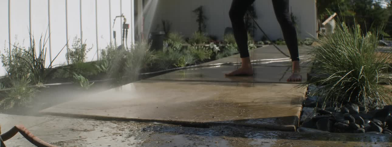 Woman stands barefoot on sidewalk in front yard and washes it with a hose
