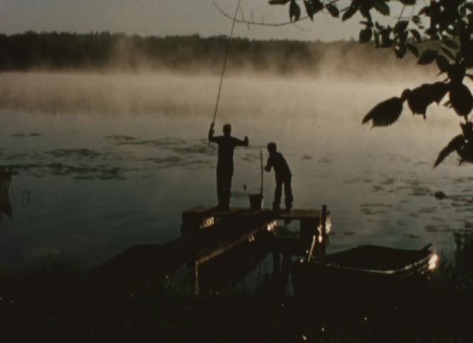 Silhouettes of two people fishing on a dock as fog rises from the water in the distance