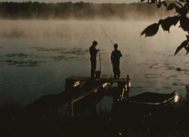 Two youth are silhouetted against foggy lake fishing off dock