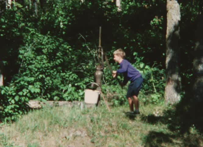 A boy operates the handle of a water pump to fill a bucket outdoors during the day