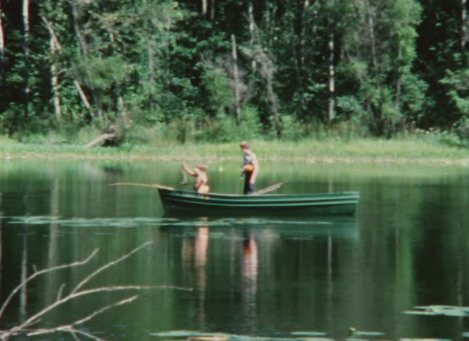 Two people are in a small green boat in the water near the woods and one is standing and the other is holding a fishing pole