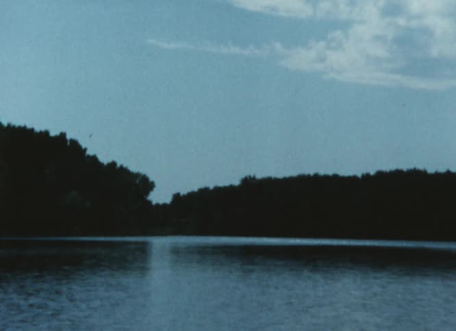 Light reflects on the ripples in the water of a lake surrounded by woods and a building in the woods