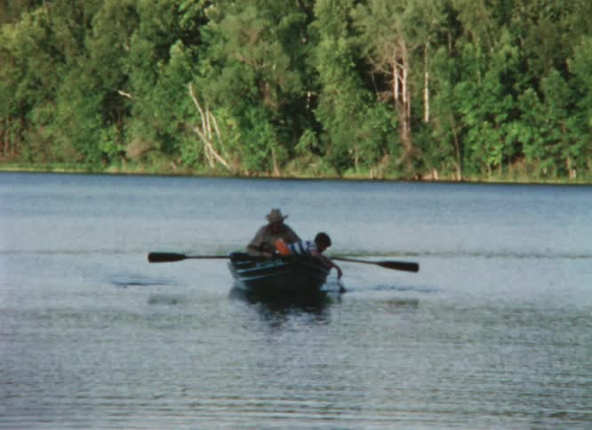 A man is rowing a small boat in the lake near woods and there is a person reaching over the side of the boat