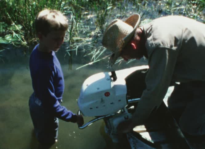 A man and a boy are at the edge of a lake looking at the motor on a small boat